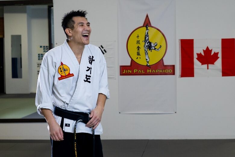 A man is laughing, wearing a white gi secured with a black belt. He's standig in a dojo, the wall behind him decorated with a Canadian flag and a poster reading 'Jin Pal Hapkido.' The same emblem is on his gi.