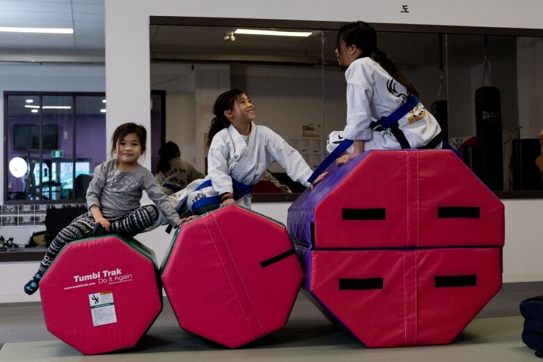 Three girls, arranged left to right from youngest to oldest, sit on three progressively larger octagonal gymnastic blocks in a dpojang. The older two are wearing white gis tied with blue belts.