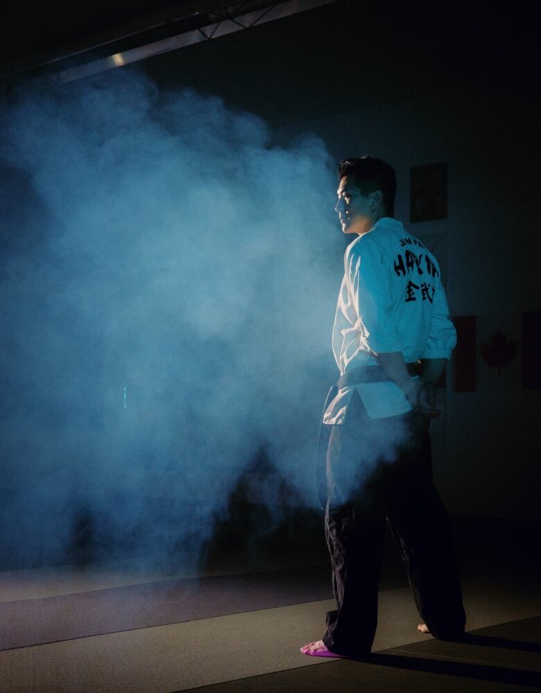 A man stands in a dark room surrounded by smoke, lit in blue tones. He is wearing black pants and a white gi.