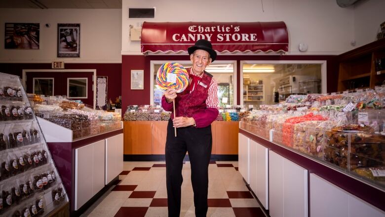 A man wearing a colourful jacket and hat holds an oversized lollipop in front of a sign that says 