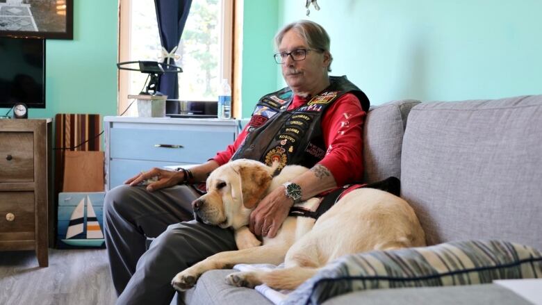 A man in a veteran's vest and a yellow lab in a service dog vest sit together on a couch. 