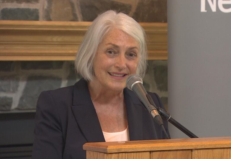 A smiling woman in a black suit stands at a lectern.