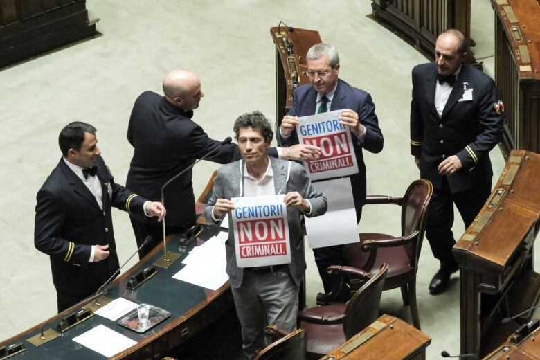 Two men in suits wear placards that read: 'Genitori! Non criminali!' which translates to 'Parents! Not criminals!' in a political chamber in Rome. They are surrounded by three men in uniform. 