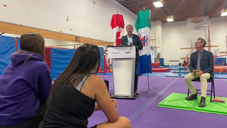 Inside a gymnasium filled with gymnastics equipment, a man in a suit at a podium speaks to a crowd.