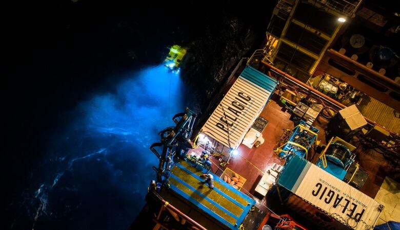 A yellow robotic vehicle going into the dark water at night. It's being lowered in attached by a cable to a ship.