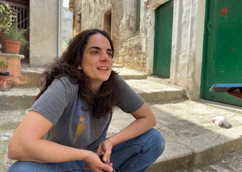 A woman with shoulder-length dark hair smiles as she talks while sitting on stone steps outdoors.