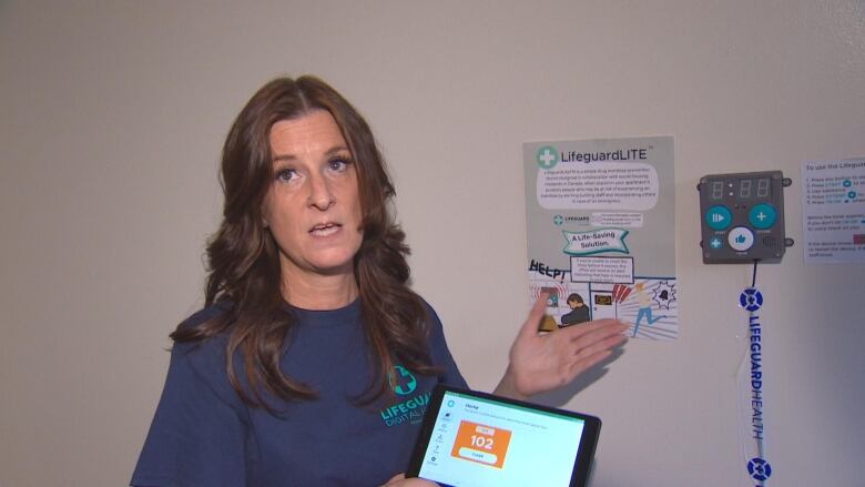 A woman with long hair holds a tablet and gestures towards a wall-mounted box with a timer on it.