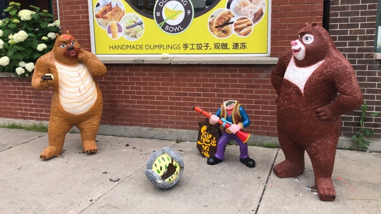 Three ceramic statues on the sidewalk. There are two bears flanking a headless man. His head sits in the foreground, having been torn off the statue. It has a hole in it. 