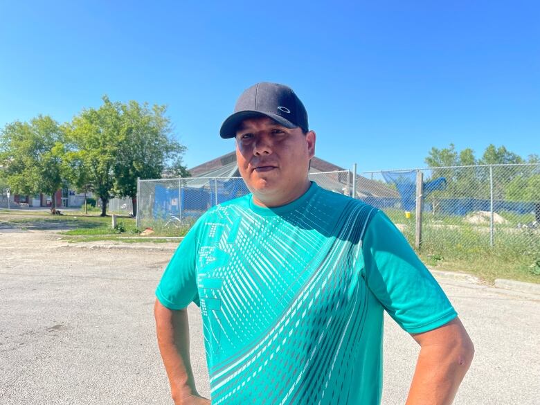 A man wearing a black cap and a teal shirt looks into the camera lens. 