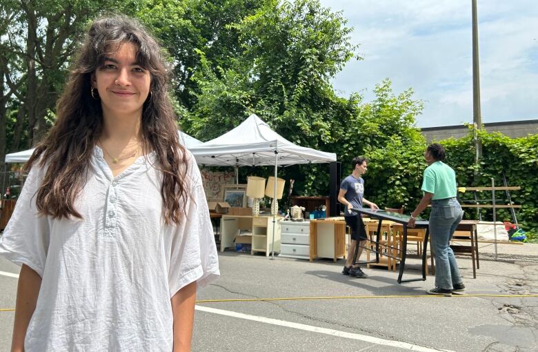 A white woman in a white t-shirt stands in front of a garage sale.