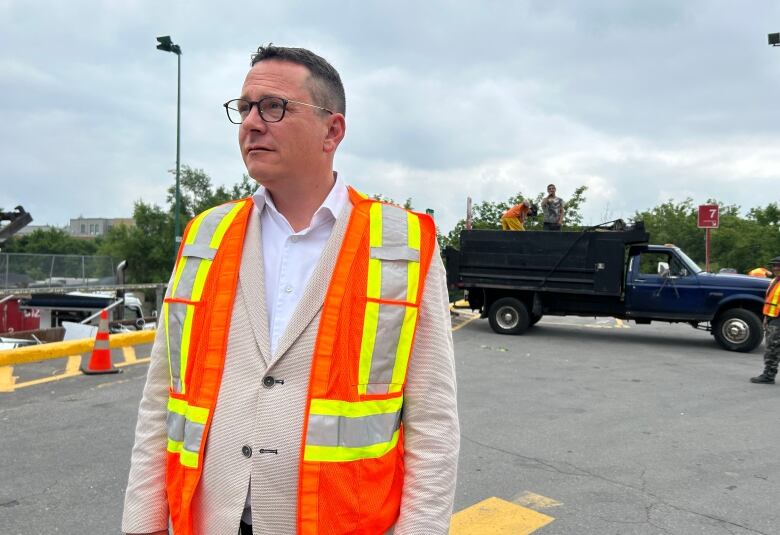 A white man wearing glasses, a pale plazer and an orange safety vest stands in a lot where people are throwing junk into containers.