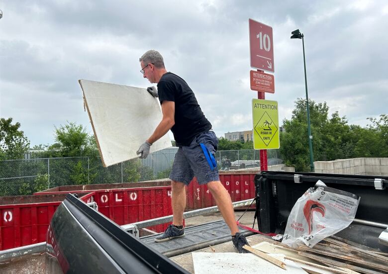 A man standing in the back of a pickup truck throws drywall into a container.