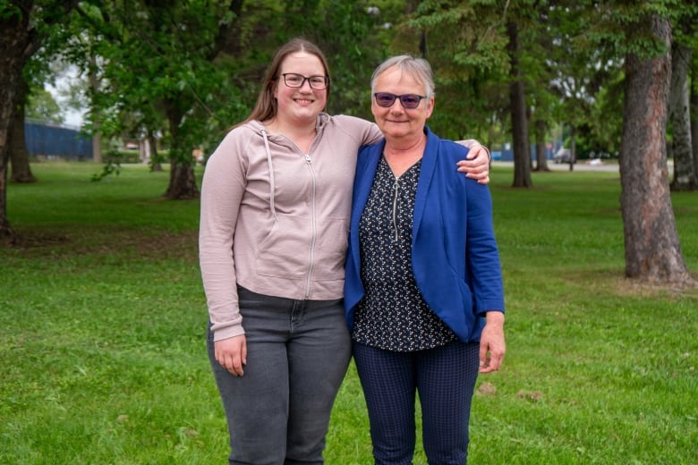 Two women stand together in a green grassy park with their arms around each other.