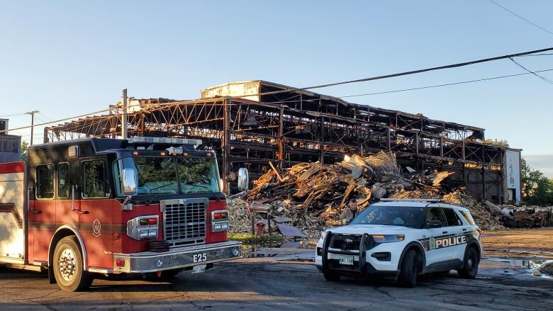 A red fire truck at the left foreground and a black-and-white police cruiser at the right foreground. In the background is the steel frame of a building that burned. All around it is rubble.