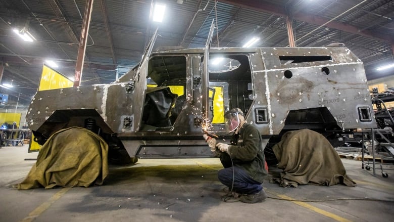 A large, uncompleted four-wheel vehicle sits on a warehouse floor, its tires shrouded in green coverings. A man wearing a protective mask holds a tool in both hands as sparks fly off it and an open door. 