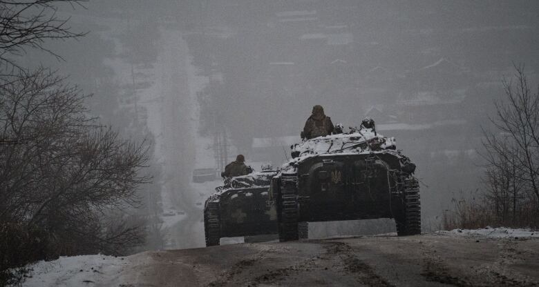 Two military vehicles move along a road with a community visible further down the road. The area and the military vehicles have snow on them.