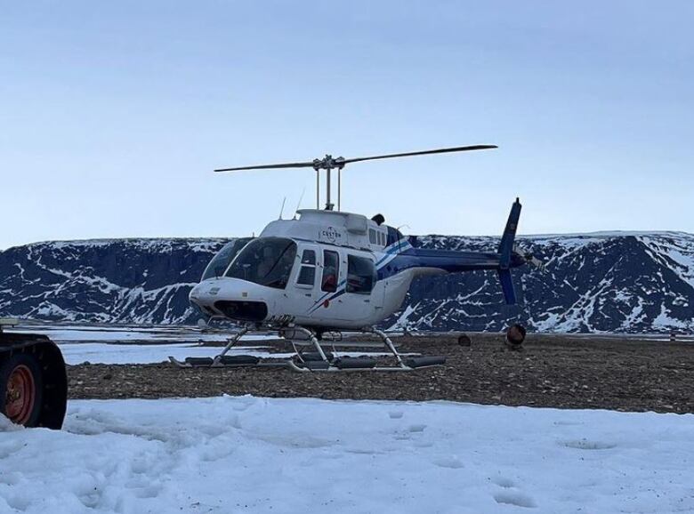 A helicopter sits on a rocky, snowy expanse, with mountains in the background.