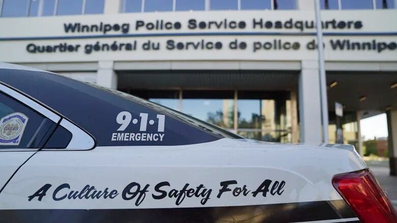 The back half of a black and white police cruiser is seen close up with a building in the background that says Winnipeg Police Service Headquarters.