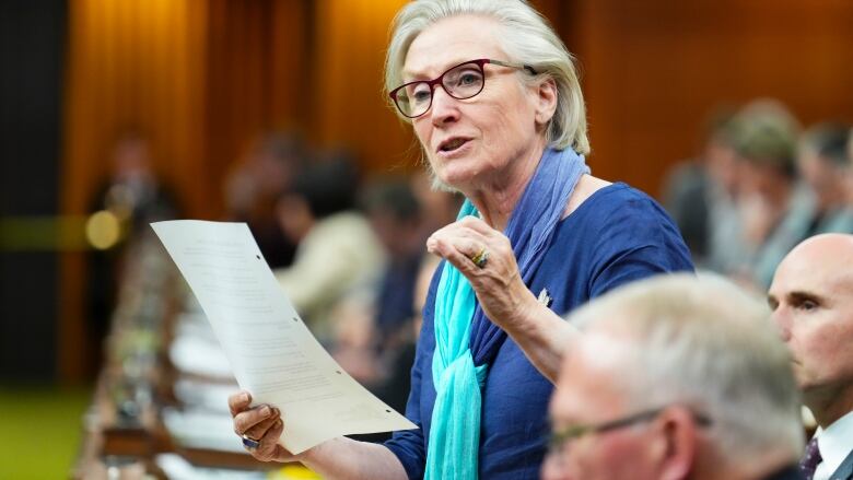 A woman wearing glasses stands in the House of Commons speaking and holding a piece of paper.