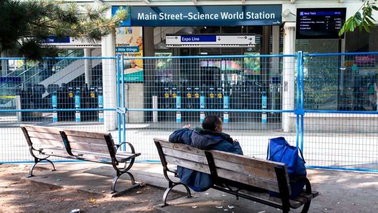 A man in a navy blue coat is pictured from the back, sitting on a bench with a blue tote bag across the Main Street-Science World SkyTrain Station.