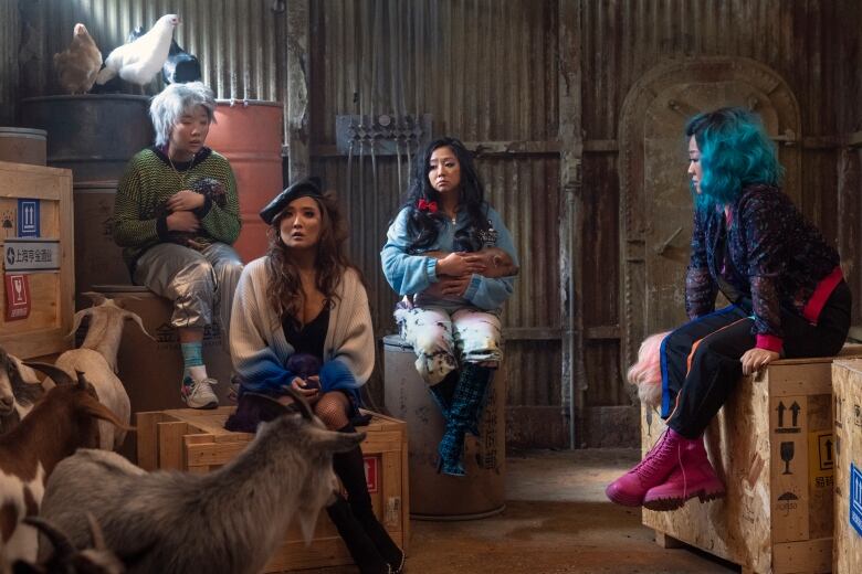 Four tired women sit on boxes in a barn. There are goats wandering nearby. 