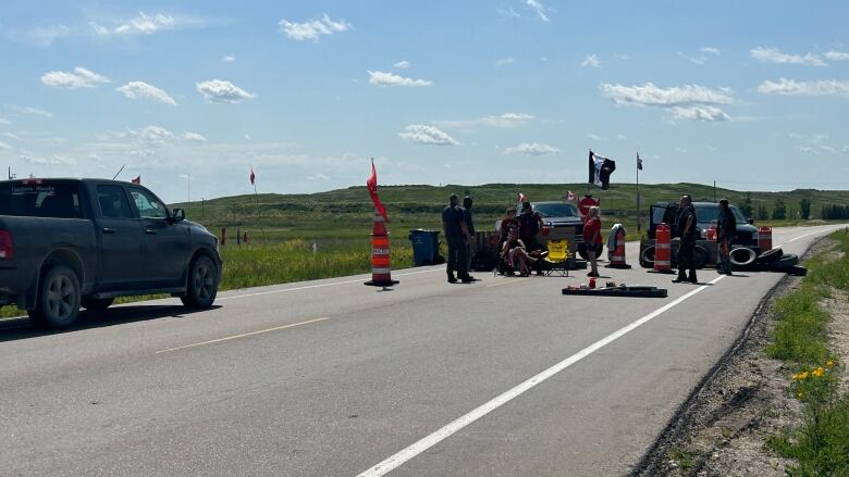 Several people stand on a road with flags, pylons, tires and vehicles park in the middle of the road.