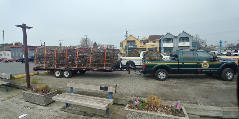 A pickup truck with official markings is towing a small trailer loaded with wire crab traps at a dock.