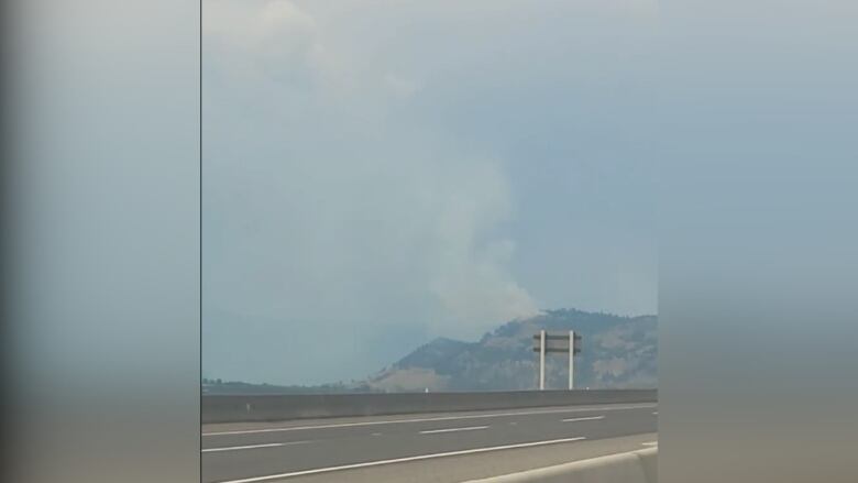 A wide shot of smoke coming out from hills behind roads with cars.