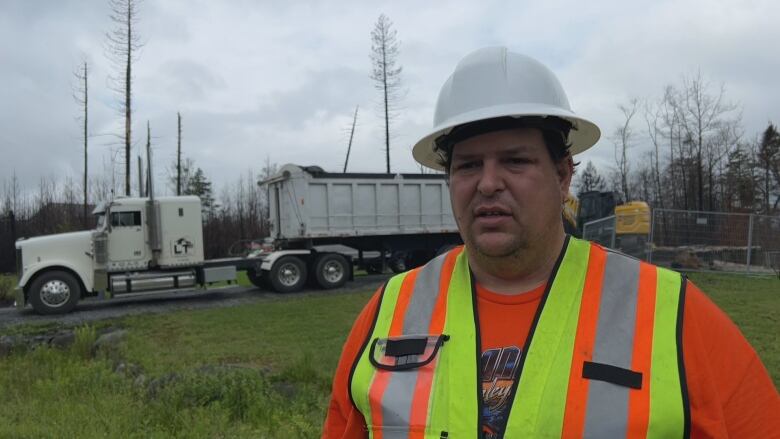 A man shown in a high visibility orange and yellow vest and a white helment is looking at the camera while a big truck and excavator works behind him.