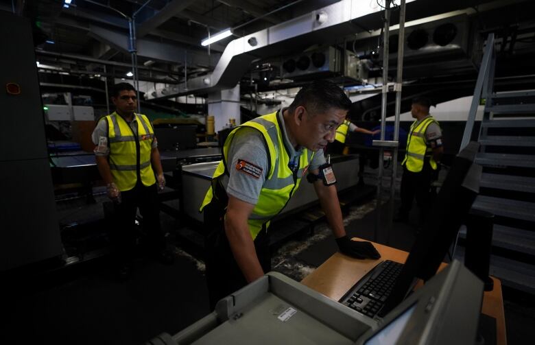 A security officer wearing a neon yellow vest looks at a computer monitor at an airport.