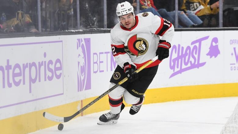 A male ice hockey payer wearing number 12 skates with the puck along the boards as fans watch from behind the glass.