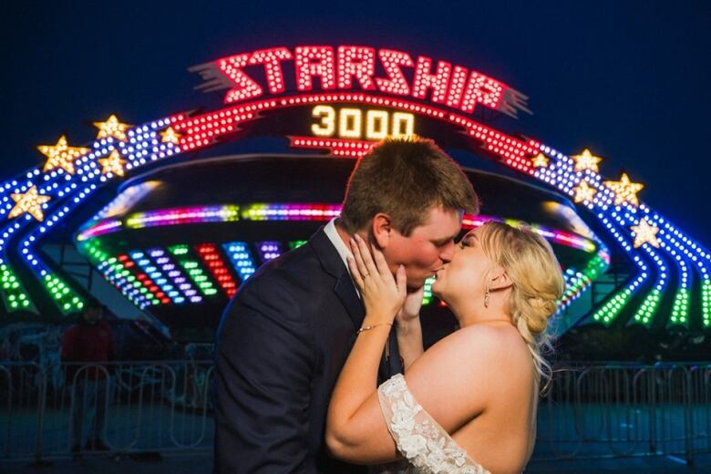 A groom and bride kiss in front of an amusement park ride. 