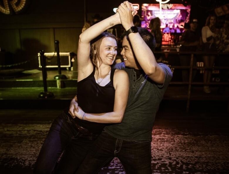 A man dances with a woman at a country dance bar, they have their hands joined and raised above their heads as they lean to one side.