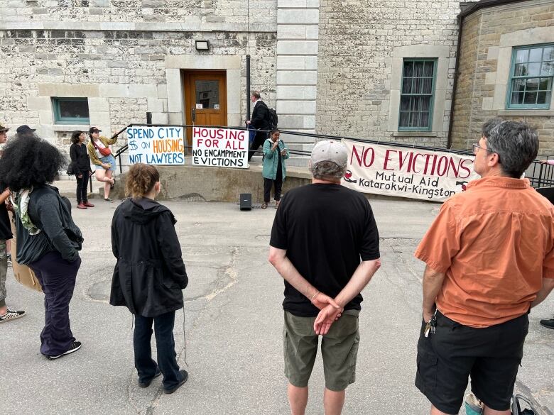 A circle of people stand in a parking lot in front of a grey stone building. There are signs on a stairway behind them saying Spend city $ on housing, not lawyers