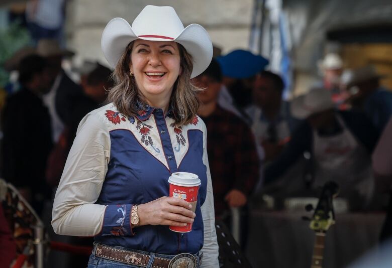 A smiling woman wearing a cowboy hat, western shirt and jeans. She is holding a coffee.