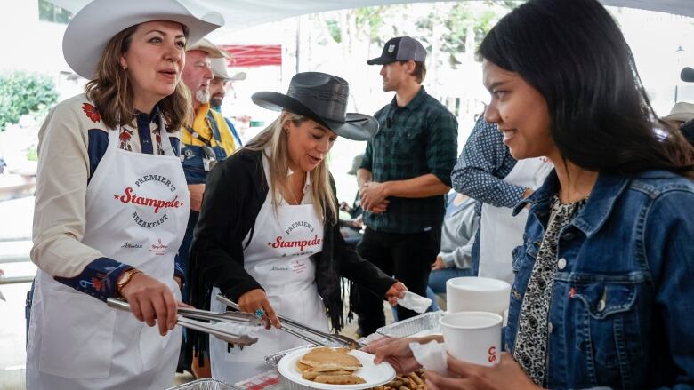 A woman holding tongs over a tray of pancakes speaks to another woman dressed in a jean jacket and holding a plate.