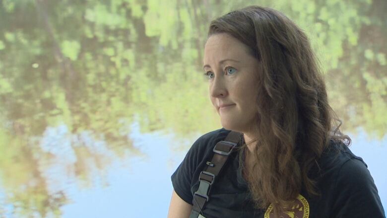 A woman wearing hip-waders looks at someone outside of the frame. She stands at the bank of the Nashwaak river.