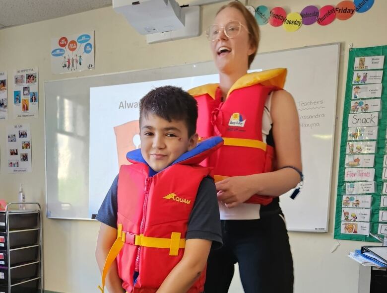 A woman wearing a life jacket stands behind a young boy who is also wearing a life jacket.