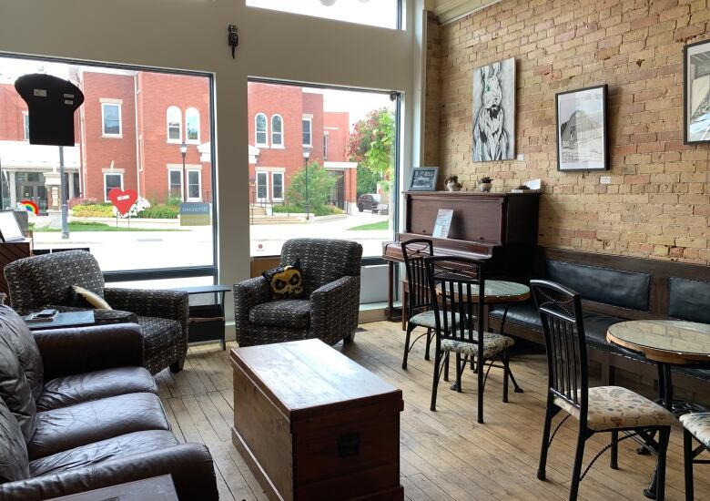 Indoor shot of a cafe, with a brick wall and piano. Numerous, high-back tables. A wooden coffee and leather couches. Two large windows. 