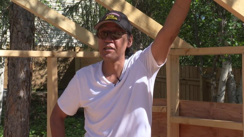 A man in white shirt and sunglasses has arm lifted up, holding wooden tent frame.