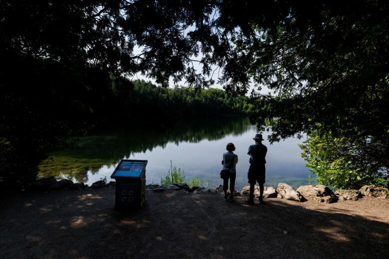 Two people stand on the shore of a lake, framed by trees, with an interpretive sign in the foreground.