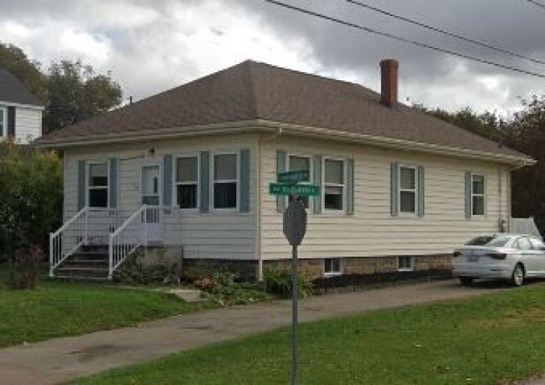 A small yellow house with a brown roof and foundation. It is situated near a stop sign and a silver car is in the driveway in front of a small white shed