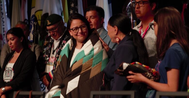 A woman is wrapped in a starblanket on a stage with other people.