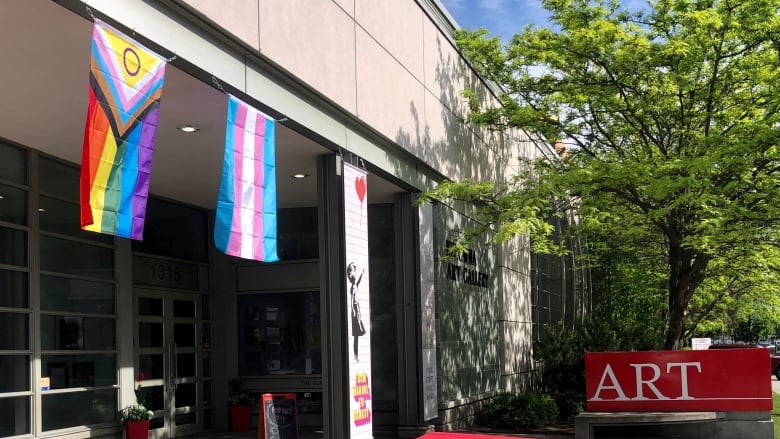 An art gallery showing a pride and trans pride flag hanging from a concrete roof over an entranceway.