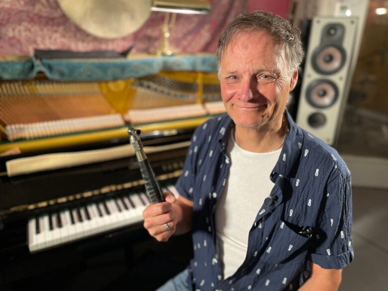 A smiling man sits in front of a piano holding a piano tuning wrench.