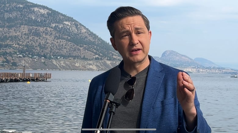 A man in a grey t-shirt and blue blazer speaks into a microphone. In the background is a lake and hills.