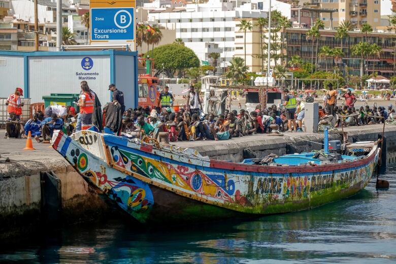 People sit on a concrete pier with a colourful wooden boat resting on the water below. 