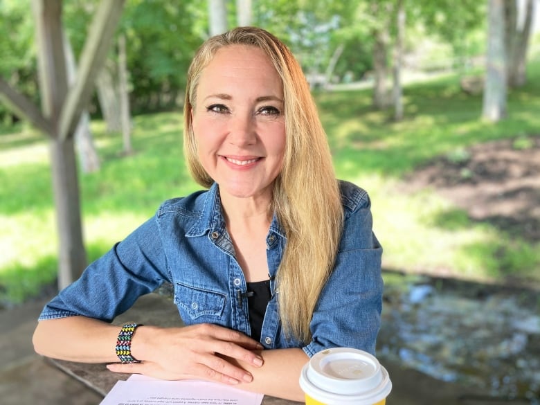 A woman with long blonde hair sitting outside at a table with her arms folded over a piece of paper. A yellow disposable coffee cup with a white lid sits on the table in front of her.