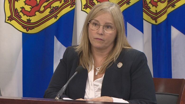 A woman with glasses sits at a table with flags behind her.