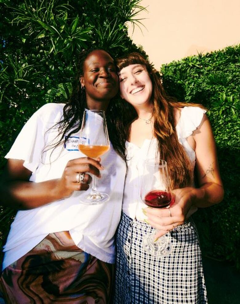 Two women stand in front of a hedge holding glasses of wine. 